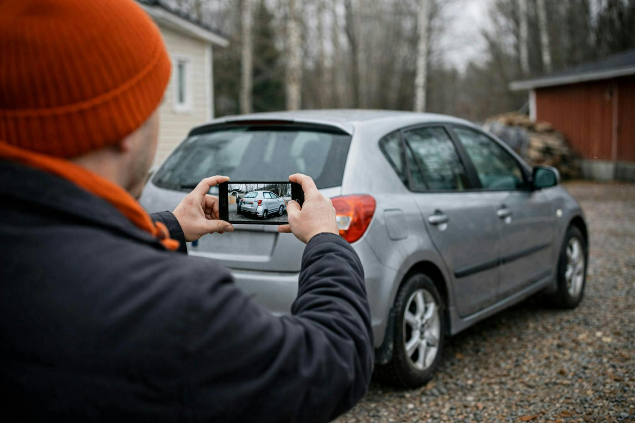 oranssipipoinen mies kuvaa kännykällä omaa renault-henkilöautoaan pihamaalla