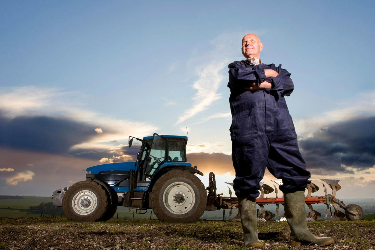 Mies seisoo kädet puuskassa haalareissa pellolla takanaan sininen traktori