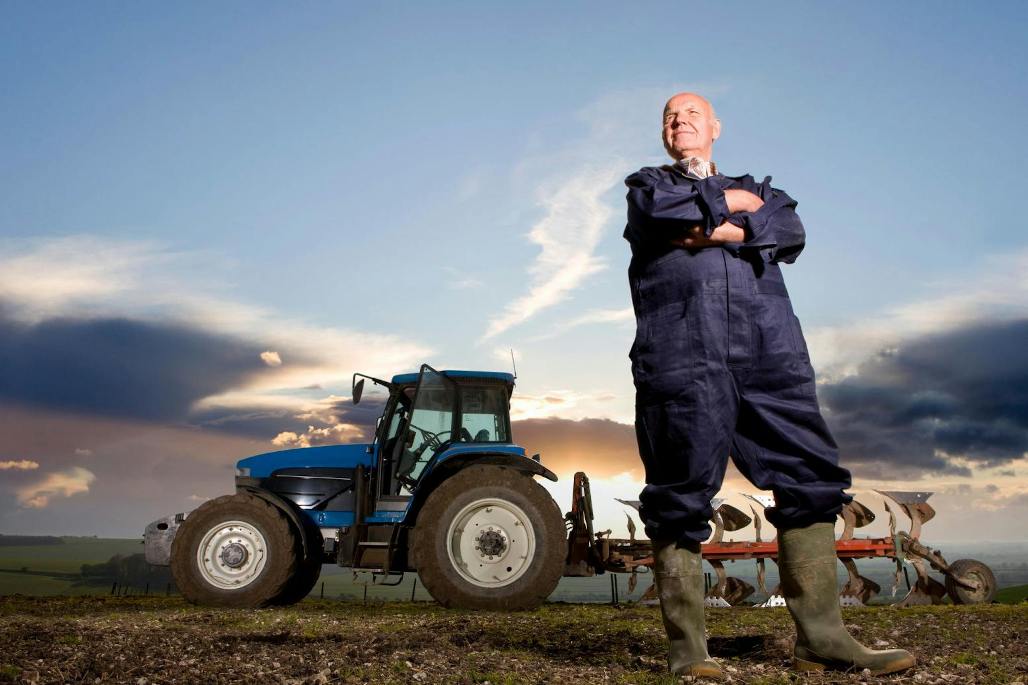 Mies seisoo kädet puuskassa haalareissa pellolla takanaan sininen traktori